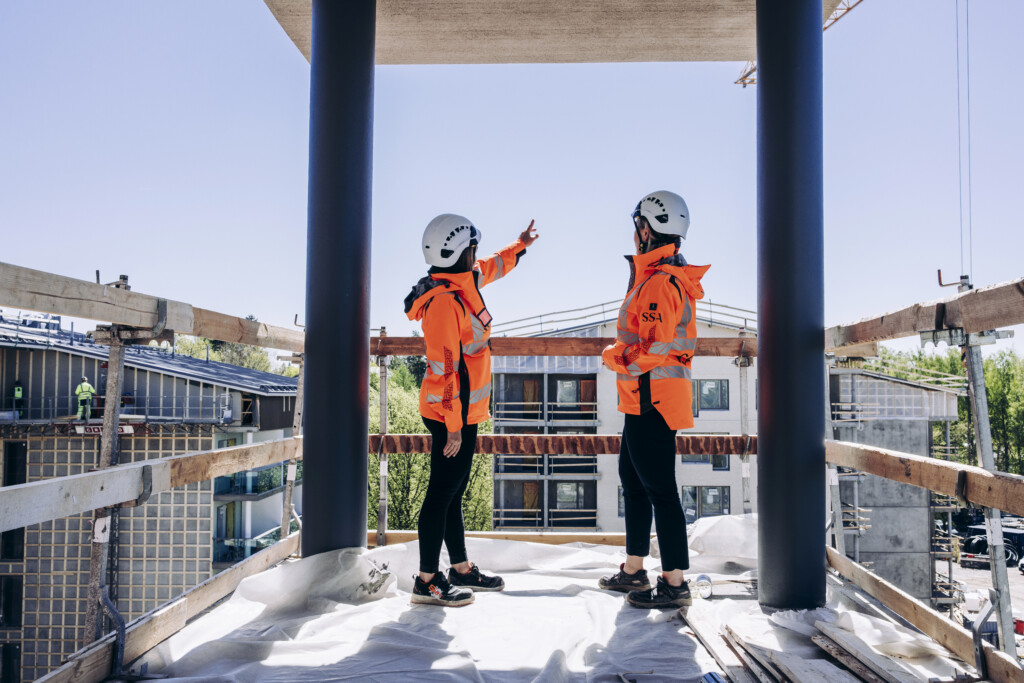 2 SSA Rakennuksen työntekijää seisovat työmaalla oranssit huomiliivit ja suojakypärät yllään